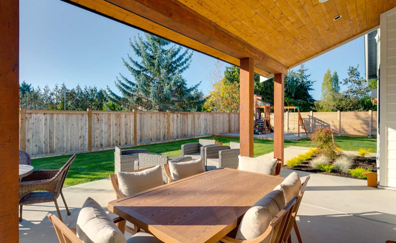 Covered backyard patio with wooden beams, seating and a table in a landscaped yard.