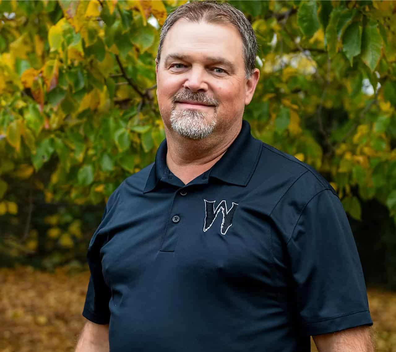 Portrait of a smiling man in a navy polo with a W logo, outdoors in autumn colors.