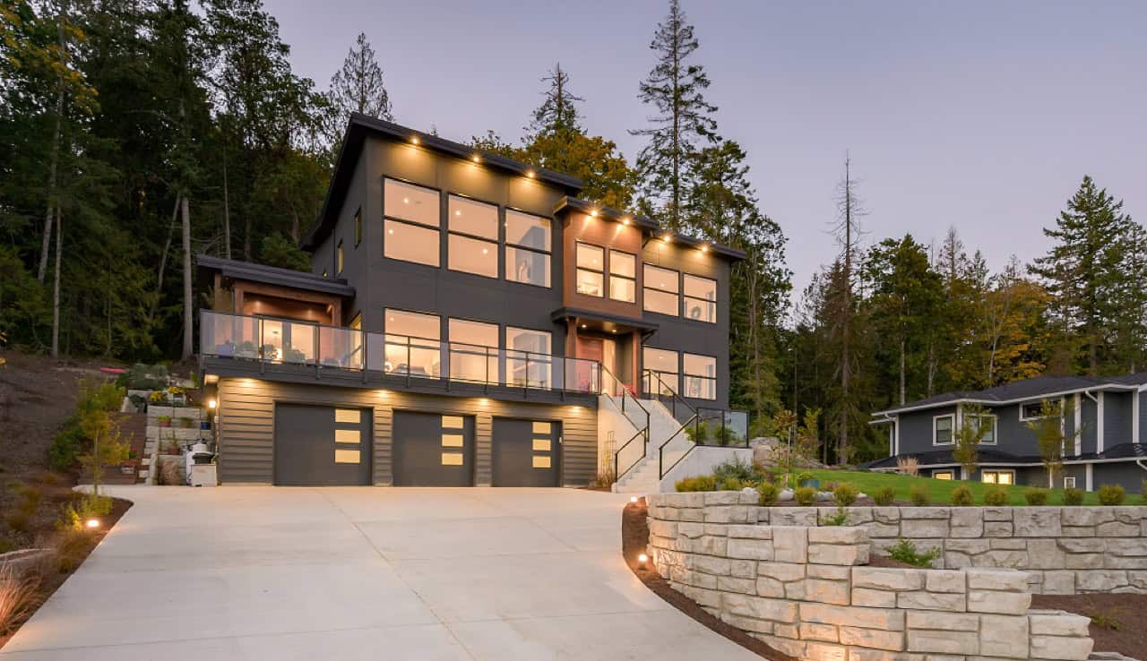 Modern custom home by White Wolf Homes in Victoria BC at dusk with multi-car garage and stone wall.