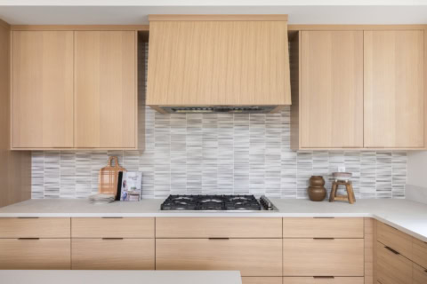 Bright modern kitchen with light wood cabinets and gas stove under a range hood.
