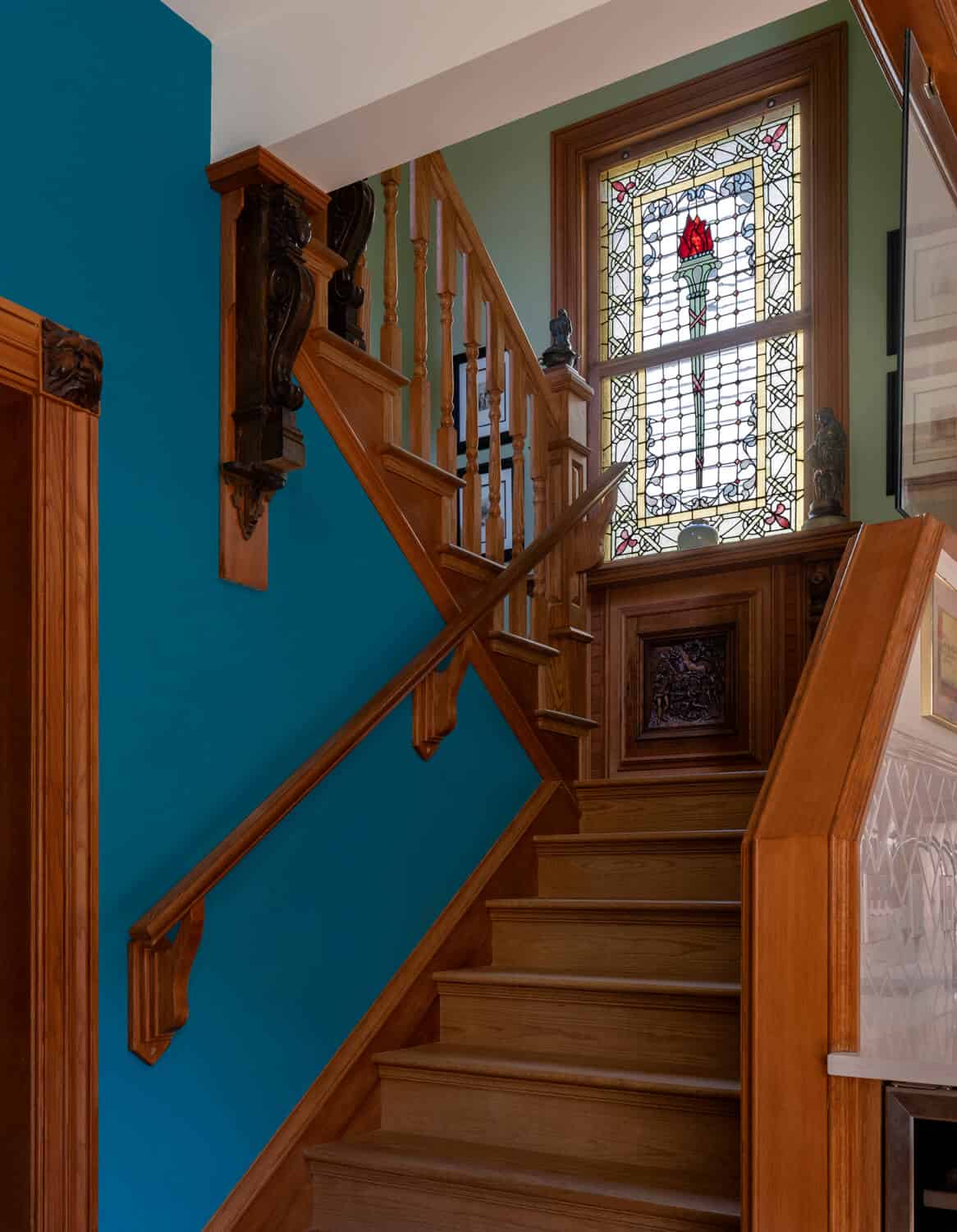 Interior staircase with teal wall, wooden banisters, and stained glass window at top.