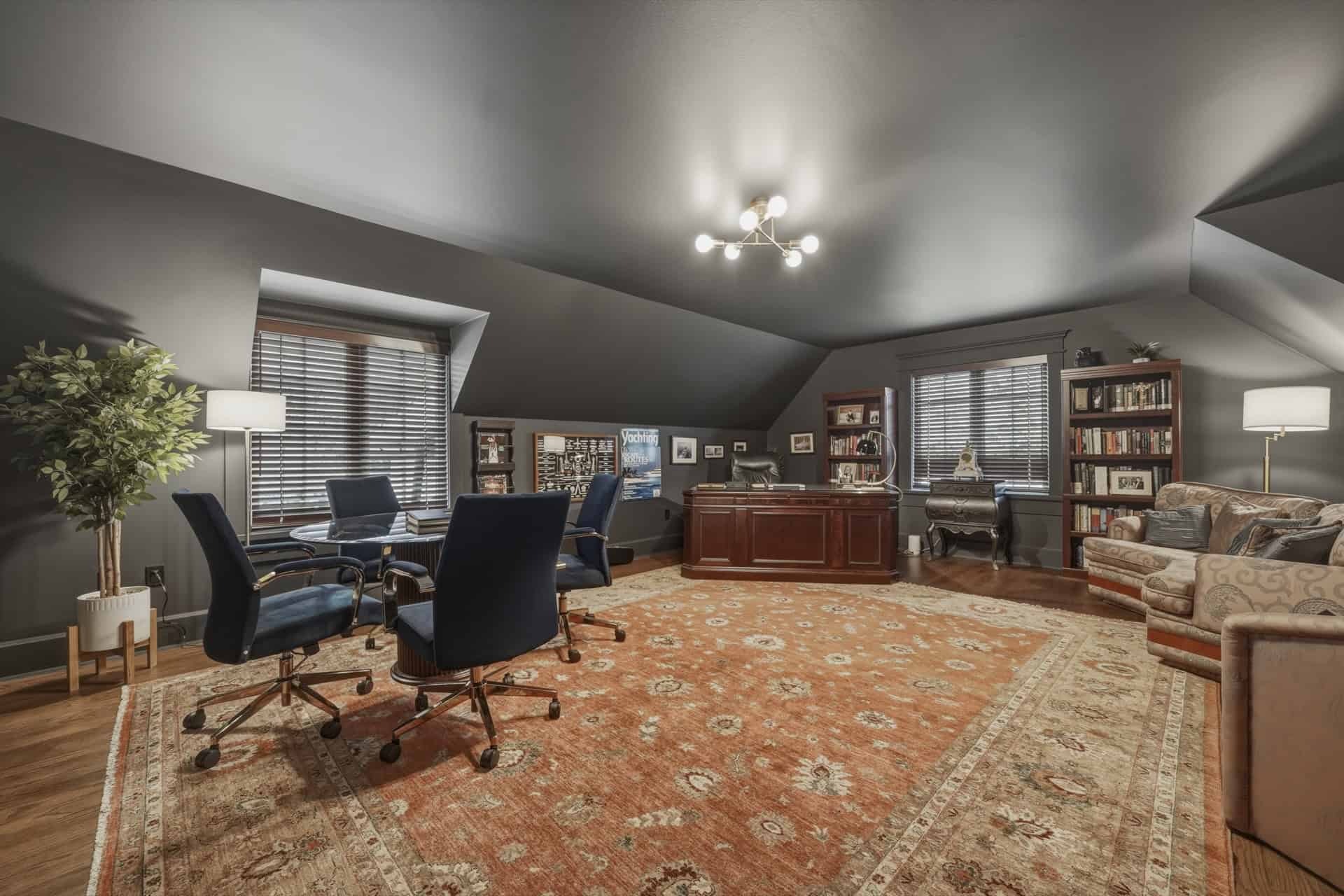 Interior of a luxury home office with dark walls, wooden desk, seating, bookshelves and warm lighting.