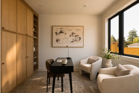 Interior view of a modern home office with beige seating, wooden cabinets, and a large window onto the garden.