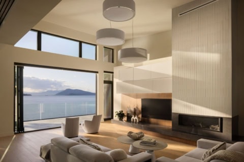 Open living room with large windows overlooking water and a distant island, with a modern fireplace.