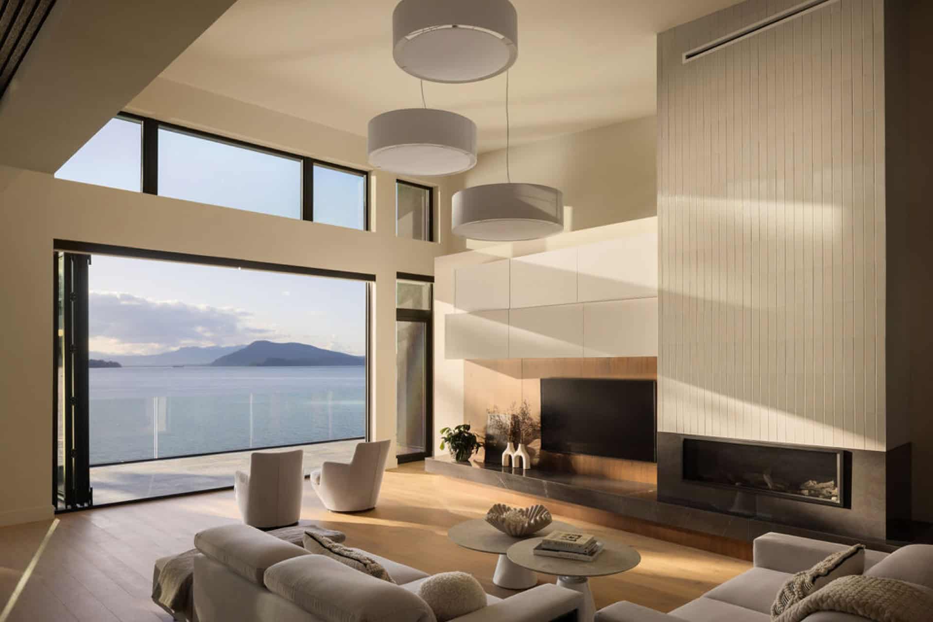 Open living room with large windows overlooking water and a distant island, with a modern fireplace.