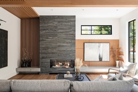 Modern living room with gray stone fireplace, wood accents, and large windows.