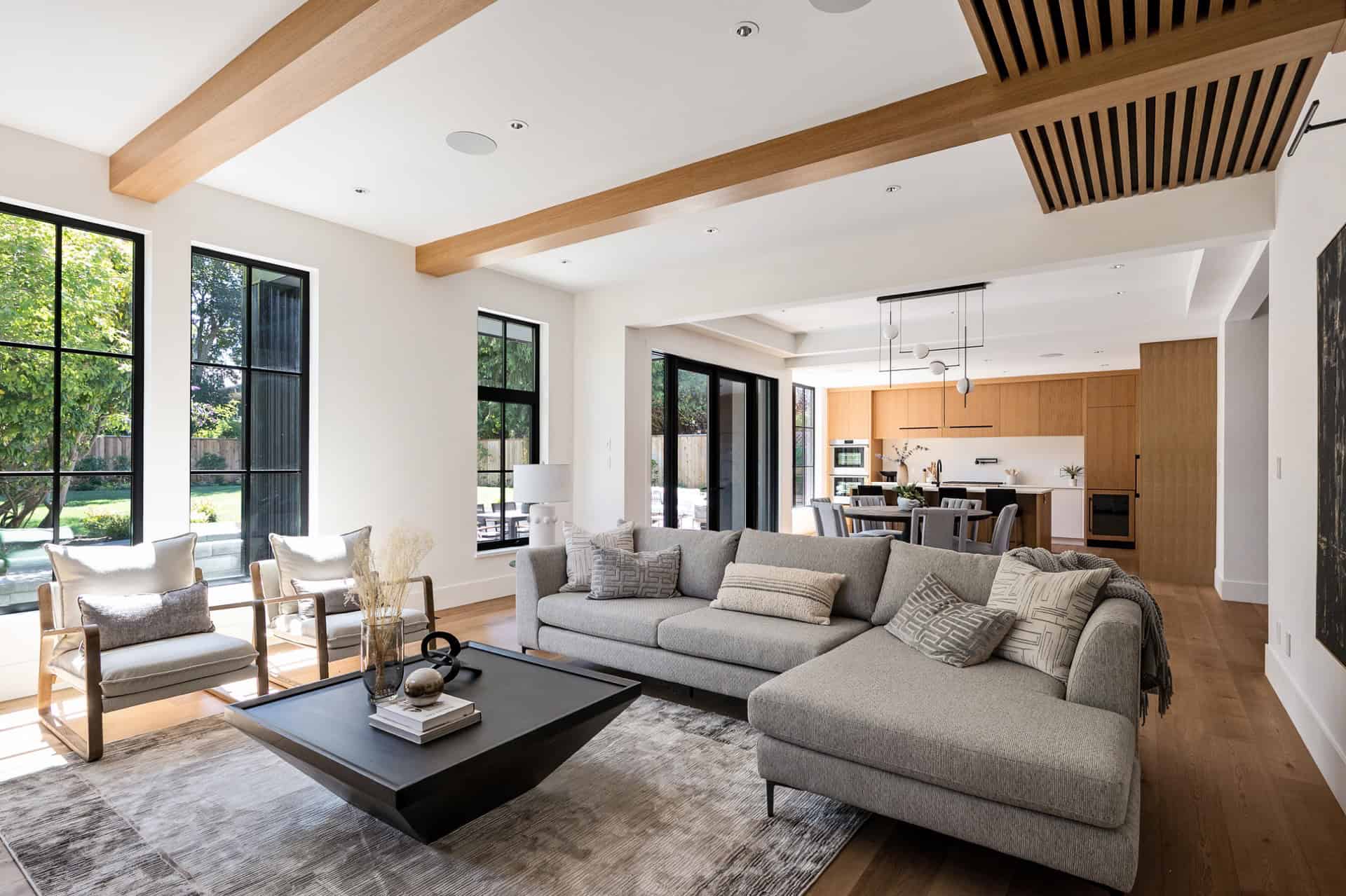 Open-plan living room with large windows, gray sofa, wooden beams, and contemporary kitchen in the background.