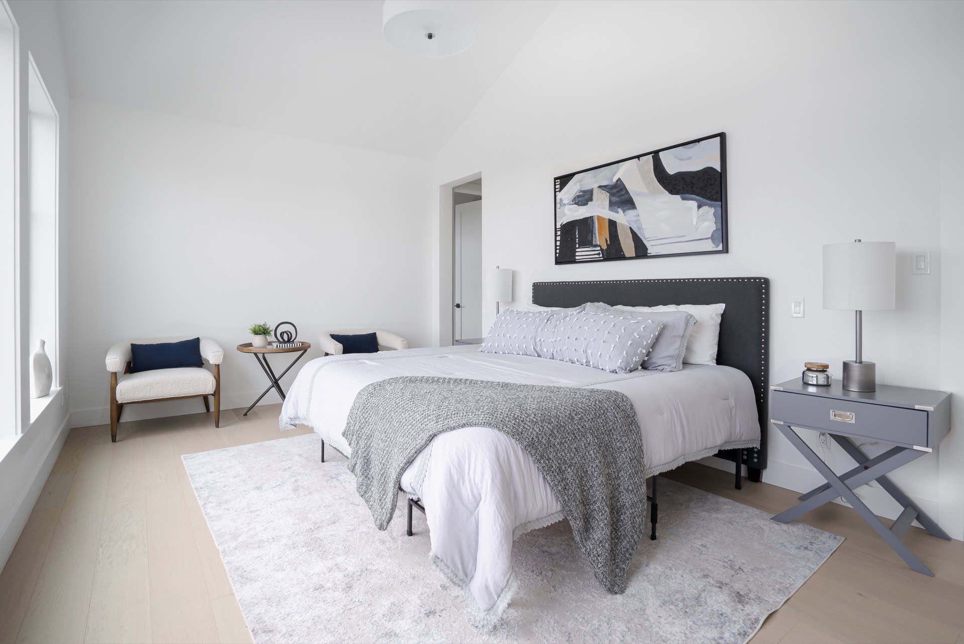 Bright modern bedroom with a gray upholstered bed, matching nightstands, and abstract wall art.