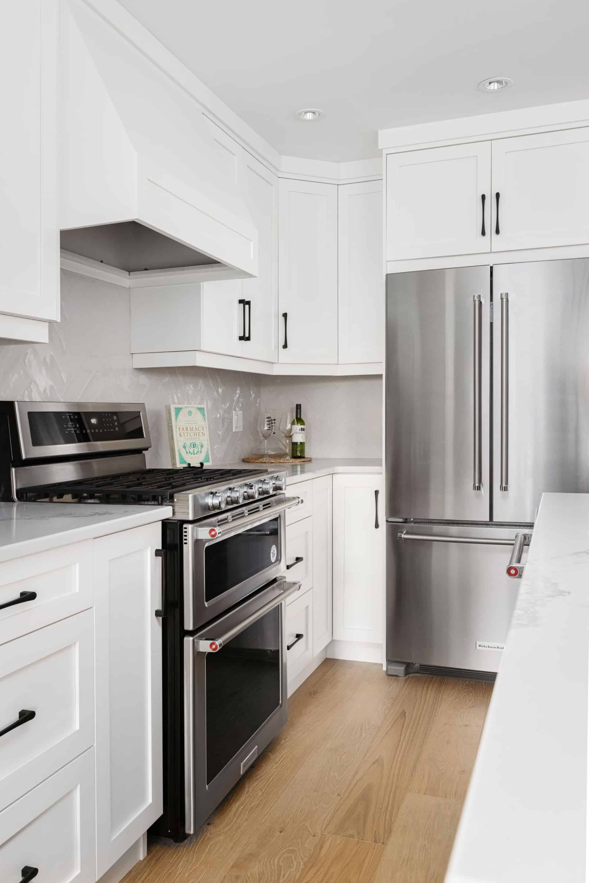 Modern white kitchen with stainless steel fridge and range, light wood floor.