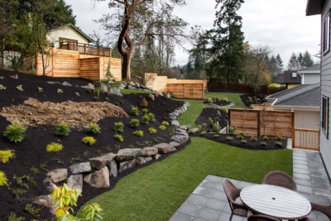 Backyard landscape with tiered rock retaining walls, fresh mulch, and seating area under a wooden structure.