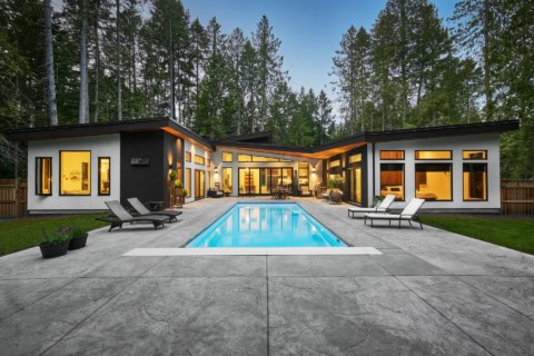 Modern single-story home with illuminated windows and a rectangular pool in a wooded setting.