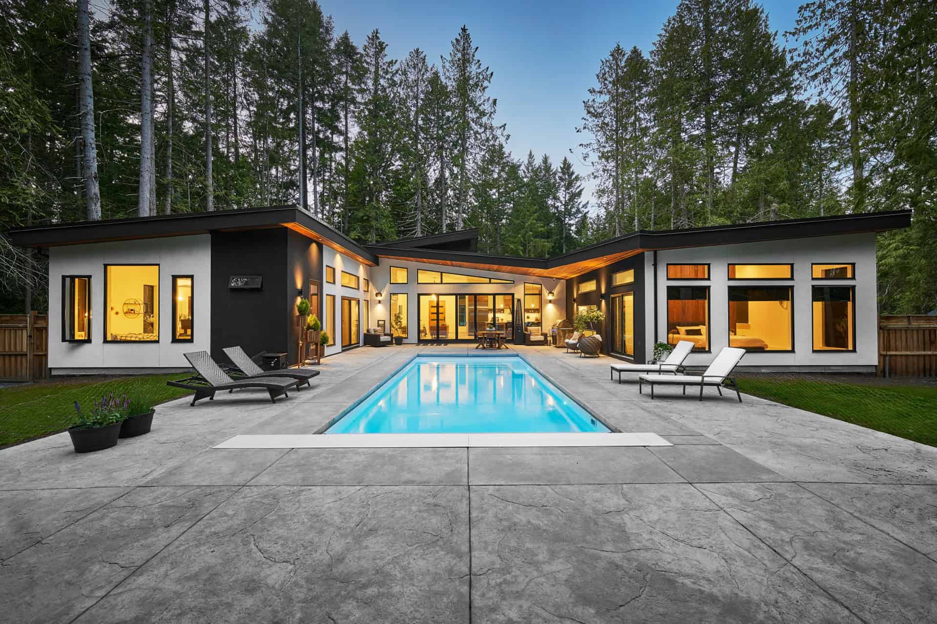 Modern single-story home with illuminated windows and a rectangular pool in a wooded setting.