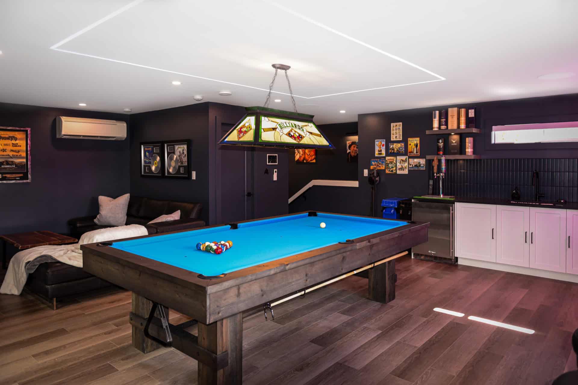 Modern game room with a blue pool table and neon lighting, sleek dark walls, and a kitchenette.