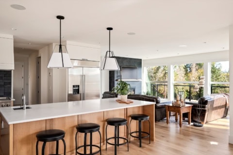 Open-concept kitchen island with four stools and a bright living area with large windows in a modern home.