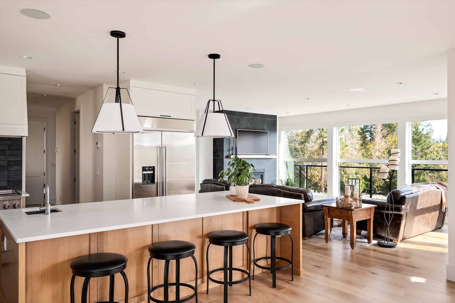 Open-concept kitchen island with four stools and a bright living area with large windows in a modern home.