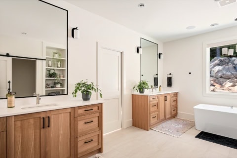Bright modern bathroom with dual wooden vanities, wide mirrors, potted plants, and a freestanding soaker tub.